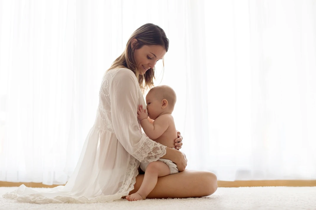 A mother in a white garment breastfeeding her baby while sitting on a soft surface with bright curtains in the background.